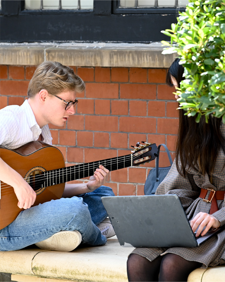 Two students playing the guitar and working on a computer, sitting on a bench.